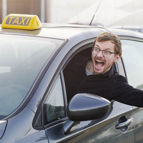 Un hombre con gafas, visiblemente emocionado o sorprendido, asoma la cabeza y el brazo por la ventana de un taxi. El coche tiene un letrero amarillo de 'TAXI' en el techo, y el hombre parece estar gritando o gesticulando con entusiasmo mientras mira hacia afuera.