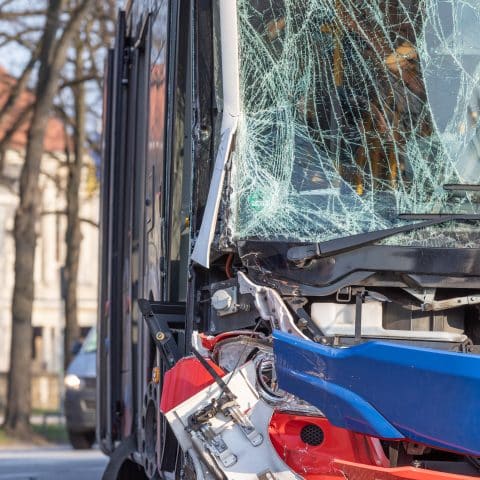 Autobús dañado con el parabrisas roto y la parte delantera destrozada tras un accidente. El cristal está visiblemente agrietado, y el frente del vehículo muestra signos de colisión grave. La imagen está tomada en una calle con árboles y edificios de fondo.