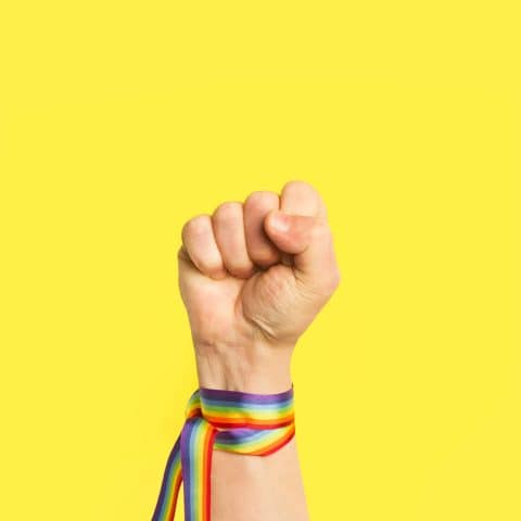 Puño levantado con pulsera de bandera arcoíris LGBT sobre un fondo amarillo brillante liso y aislado. Vista de frente y de cerca