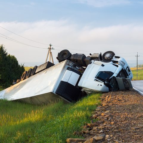 Camión volcado en el arcén de una carretera rural, con la cabina y el remolque completamente fuera de la vía. El accidente ocurrió en una curva, y un coche se aleja por la carretera. El paisaje circundante es verde con colinas y postes eléctricos visibles.