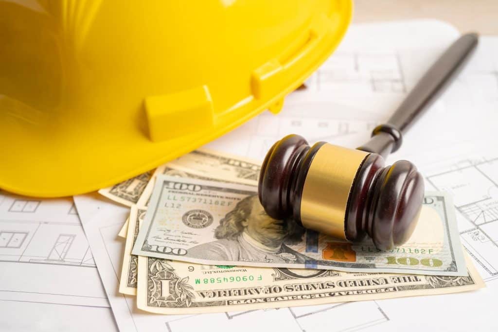Casco de seguridad amarillo, mazo de juez de madera con detalles dorados y billetes de dólares sobre planos de construcción. La imagen representa la relación entre la legislación, la industria de la construcción y aspectos financieros, como disputas legales o regulaciones en proyectos de construcción.