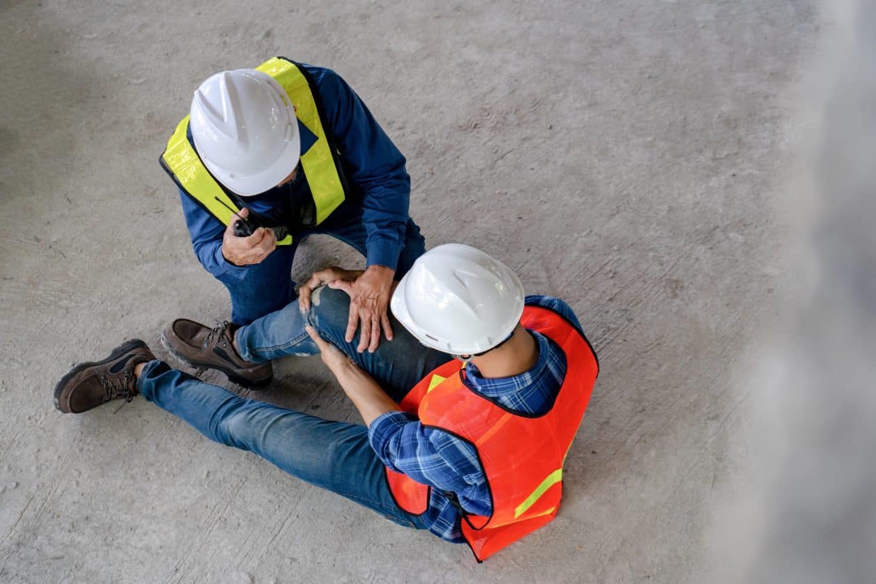 Dos trabajadores de construcción con cascos y chalecos de seguridad, uno de ellos sentado en el suelo sosteniendo su rodilla en señal de dolor, mientras el otro se inclina hacia él, usando un walkie-talkie. La escena parece representar un accidente laboral en un sitio de construcción.