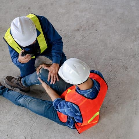 Dos trabajadores de construcción con cascos y chalecos de seguridad, uno de ellos sentado en el suelo sosteniendo su rodilla en señal de dolor, mientras el otro se inclina hacia él, usando un walkie-talkie. La escena parece representar un accidente laboral en un sitio de construcción.