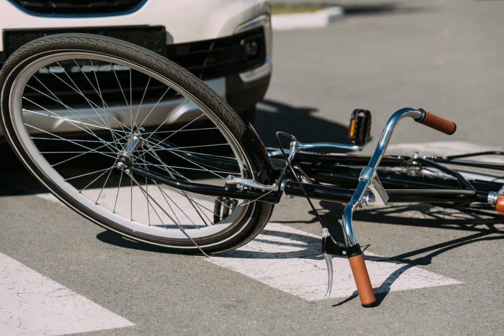 Una bicicleta tirada en el suelo frente al parachoques de un automóvil, sugiriendo un posible accidente. La rueda delantera de la bicicleta está frente al coche y las marcas de un paso de peatones son visibles debajo de la bicicleta. La escena ocurre en una calle pavimentada.