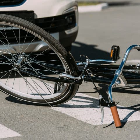 Una bicicleta tirada en el suelo frente al parachoques de un automóvil, sugiriendo un posible accidente. La rueda delantera de la bicicleta está frente al coche y las marcas de un paso de peatones son visibles debajo de la bicicleta. La escena ocurre en una calle pavimentada.